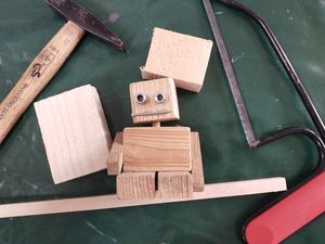 Eine aus Holzklötzen zusammen gesetzte Figur mit Augen, daneben ein Hammer und eine Zange