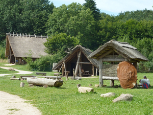 Rekonstruierte steinzeitliche Gebäude mit Holz- und Reetdächern, umgeben von grünen Wiesen und Bäumen