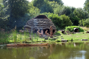 Reetgedeckte Rundhütte mit mehreren Personen am Wasser, umgeben von grüner Vegetation
